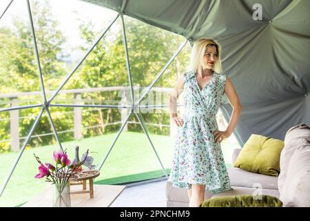 Eine Frau, die die Natur von Geo-Dome-Zelten aus betrachtet. Grün, blau, orangefarbener Hintergrund. Gemütlich, Camping, Glamping, Urlaub, Urlaub Lifestyle Konzept Stockfoto