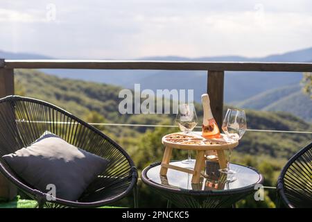 Champagner- und Cocktailgläser auf dem Glastisch auf der Außenterrasse mit Blick auf die Berge bei Sonnenuntergang Stockfoto