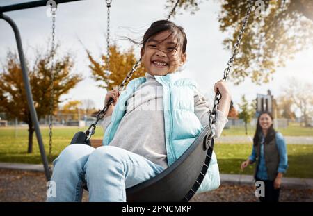 Spielen ist so wichtig für Kinder. Porträt eines kleinen Mädchens, das auf einer Schaukel im Park spielt, mit ihrer Mutter im Hintergrund. Stockfoto