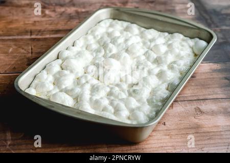 Hausgemachter, roher Focaccia-Teig, der in einer rechteckigen Metallpfanne auf Holzhintergrund ruht. Stockfoto