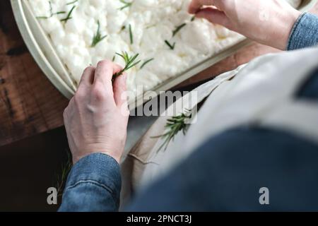 Weibliche Hände, die Rosmarin-Blätter kneifen und mit Teig Focaccia garnieren. Der Kochvorgang: Italienisches Fladenbrot Focaccia. Stockfoto