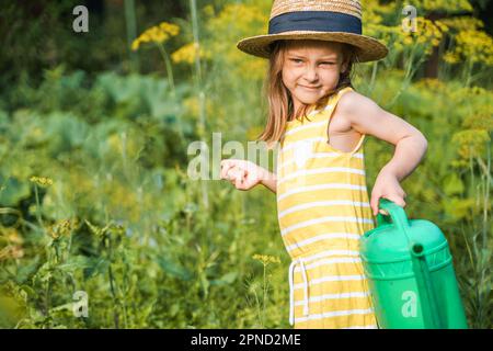 Bauernmädchen in gelbem Kleid. Kleiner Gemüsegärtner, der im Garten anbaut. Große grüne Gießkanne für frische Gurken Setzlinge. Erntehilfe arbeiten. Stockfoto