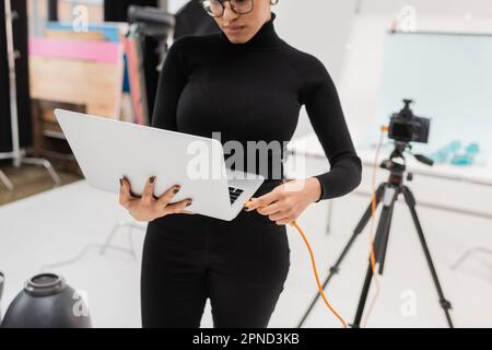 afroamerikanischer Content Produzent, der ein Kabel mit einem Laptop in der Nähe einer verschwommenen Digitalkamera im Fotostudio verbindet, Stock Image Stockfoto