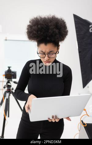 Verärgerter und verärgerter afroamerikanischer Inhaltsproduzent, der ein Notebook in der Nähe einer verschwommenen Digitalkamera im Fotostudio verwendet, Aktienbild Stockfoto