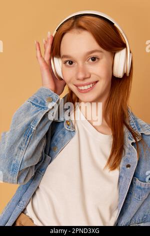 Porträt eines jungen, wunderschönen Mädchens, eines Teenagers mit roten Haaren, Kopfhörern und einem Lächeln über dem Hintergrund eines Ingwerstudios. Musik hören Stockfoto