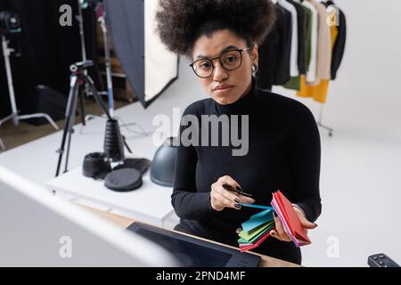 afroamerikanischer Retourenschein in schwarzem Rollkragenpulli und Brille mit Farbproben und Blick auf die Kamera in der Nähe des Grafiktabletts im Fotostudio, Stock im Stockfoto