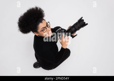 Draufsicht des überfreulichen afroamerikanischen Inhaltsproduzenten mit Brille, der eine professionelle Digitalkamera isoliert auf einem grauen Stockbild hält Stockfoto