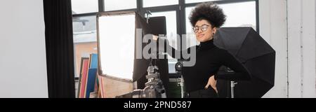 Fröhlicher afroamerikanischer Inhaltsproduzent, der mit der Hand auf der Hüfte neben dem Softbox-Reflektor posiert, im Fotostudio, Banner, Stock-Bild Stockfoto