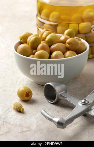 Schüssel mit grünen türkischen Oliven aus der Nähe und einem Metall-Oliven-Pitter und entsteinten Oliven davor Stockfoto