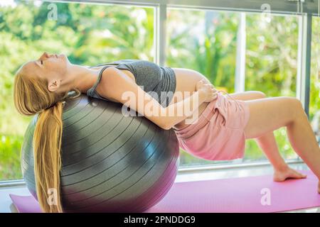 Schwangere Frau, die zu Hause auf Sportball trainiert. Schwangere Frau, die Entspannungsübungen mit einem Fitness-Pilates-Ball macht. Vor dem Hintergrund des Fensters Stockfoto