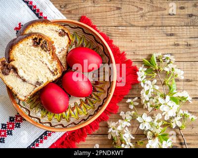 Rote Ostereier neben blühenden Zweigen und Teller mit süßem Schwammkuchen oder Kokon auf einem Holztisch Stockfoto