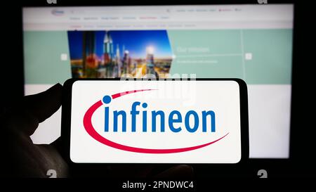 Person, die ein Mobiltelefon mit dem Logo des Halbleiterunternehmens Infineon Technologies AG auf dem Bildschirm vor der Webseite hält. Konzentrieren Sie sich auf das Display des Telefons. Stockfoto