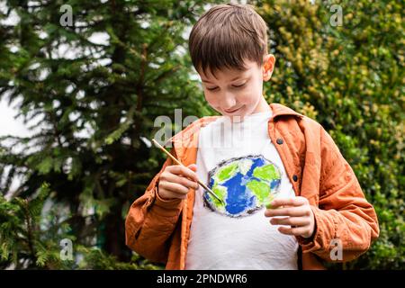 Nachhaltiges Lifestyle-Konzept. Schuljunge, der Erdkugel auf T-Shirt malt. Grüne Energie, ESG, erneuerbare Ressourcen, Umweltschutz, Schutz des Planeten Stockfoto