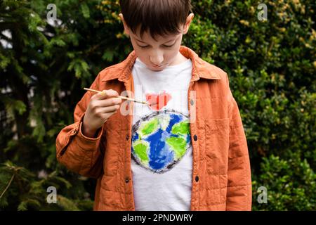 Nachhaltiges Lifestyle-Konzept. Schuljunge, der Erdkugel auf T-Shirt malt. Grüne Energie, ESG, erneuerbare Ressourcen, Umweltschutz, Schutz des Planeten Stockfoto