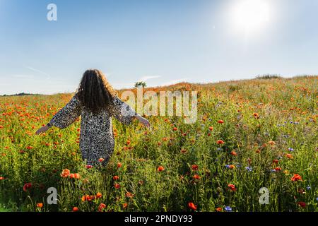 Eine lockige, langhaarige Frau in Übergröße steht an einem Sommertag inmitten eines Mohnfeldes mit seitlich gestreuten Armen. Stockfoto