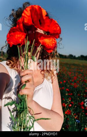 Selfie einer lockigen rothaarigen Frau, die ihr Gesicht mit einem Haufen roter Mohnblumen unter einem Mohnfeld bedeckt. Stockfoto
