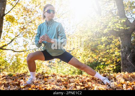 Eine junge Frau streckt sich an einem Herbsttag im Freien das Bein, trägt Uhren, Sonnenschutz und Kopfhörer. Stockfoto