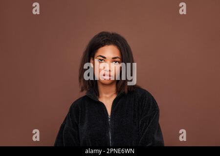 Wunderschöne junge Frau mit schwarzem Haar, die vor braunem Hintergrund in die Kamera schaut Stockfoto