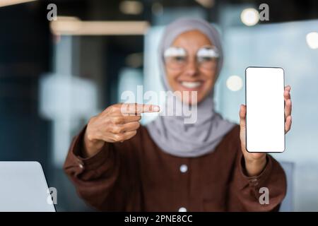 Eine junge Frau in einem Hijab mit verschwommenem Hintergrund hält ein Telefon in der Hand und lächelt. Zeigt der Kamera einen weißen Bildschirm an und zeigt mit dem Finger auf das Telefon. Stockfoto