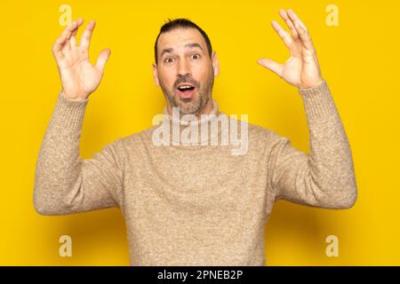 Bärtiger hispanischer Mann in seinen 40s Jahren mit beigefarbenem Rollkragenpullover, der seine Arme nach einer großen Überraschung, isoliert auf gelbem Studio, kräftig hochhob Stockfoto