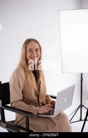 Eine Geschäftsfrau im Anzug arbeitet, tippt auf einem Laptop-Apfel und sitzt auf einem Produzentenstuhl. Porträtblonde Assistentin mit Computer MacBook. Vertikal Stockfoto