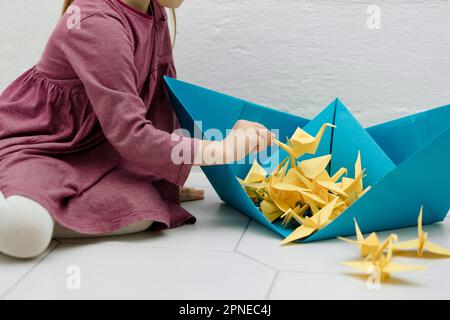 Ein kleines Mädchen spielt mit einem gelben Origami-Kran in der Hand und legt ihn in ein blaues Schiff Stockfoto