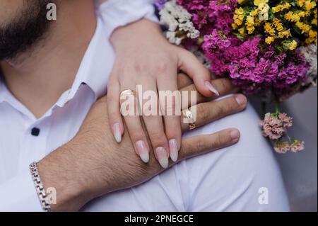 Eheringe an den Händen der Frischvermählten, ein Blumenstrauß im Hintergrund. Goldene Ringe an der Hand eines Mannes und einer Frau Stockfoto