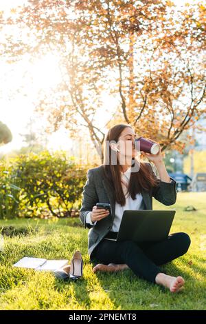 Blick auf eine barfüßige Geschäftsfrau, die mit einem Laptop auf einer Wiese sitzt und nach der Arbeit Kaffee zum Mitnehmen trinkt. Erfolgreiche Frau Stockfoto