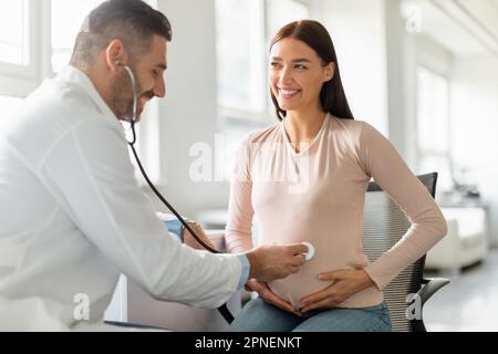 Ärztliches Leistungskonzept. Männlicher Arzt untersucht den Bauch einer schwangeren Frau mit Stethoskop, hört dem Herzschlag des Babys zu Stockfoto