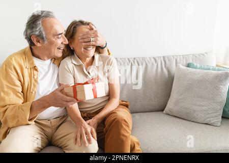 Liebevoller, glücklicher älterer Ehemann, der die Augen seiner Frau schließt, Geschenkbox präsentiert, Begrüßung mit Geburtstag oder Hochzeitstag, freier Platz Stockfoto