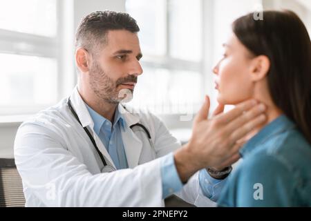 Freundlicher männlicher Arzt mittleren Alters, der Halsschmerzen oder Schilddrüsen prüft, den Hals der jungen Patientin berührt Stockfoto