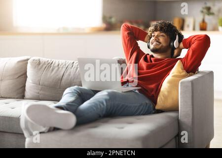 Junger Indischer Mann Mit Kabellosen Kopfhörern, Der Sich Auf Dem Sofa Mit Einem Laptop-Computer Entspannen Kann Stockfoto