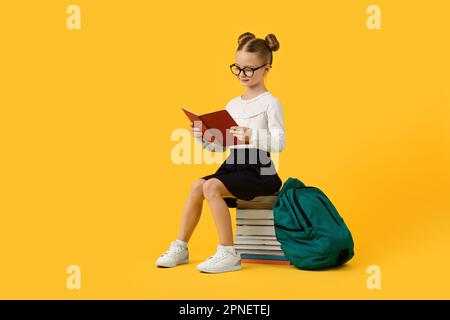 Süßes kleines Schulmädchen, das auf dem Bücherstapel sitzt und die Aufgabe im Arbeitsbuch liest Stockfoto