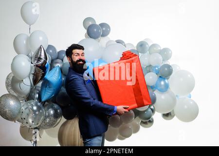 Geburtstagsgeschenk. Glücklicher bärtiger Mann mit Heliumballons und einer großen Geschenkbox. Geburtstagsparty. Geschäftsmann im Anzug mit großer Geschenkbox. Glücklich Stockfoto