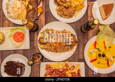 Ein Set peruanischer Gerichte mit Papas a la huancaina im Zentrum, ein Quesillo, Steaks mit Reis und Kochbananen, Arros Chaufa und Getränke auf einem braunen Stockfoto