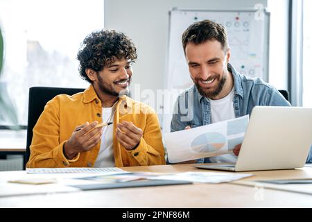 Zwei fröhliche männliche Mitarbeiter aus verschiedenen Rassen sitzen am Tisch im Büro, schauen sich den Laptop-Bildschirm und die Dokumente an, arbeiten an einem gemeinsamen Projekt, beraten sich, diskutieren Ideen und Strategien, bewerten Risiken Stockfoto