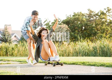 Ein Mann schubst eine Frau, die im Park auf dem Longboard sitzt Stockfoto