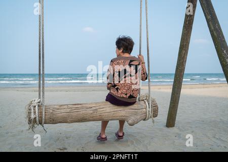 Rückblick auf eine ältere Frau, die auf einer Holzplanke mit Blick auf das Meer sitzt. Pensions- oder Urlaubskonzept. Stockfoto