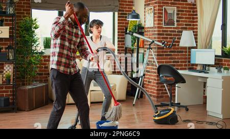 Ein junges Paar, das Hausarbeiten macht und singt, Musik für Frühjahrsputz hört. Fröhliche Freundin und Freund, die Fußböden fegen, Staubsauger benutzen, im Wohnzimmer tanzen. Stockfoto