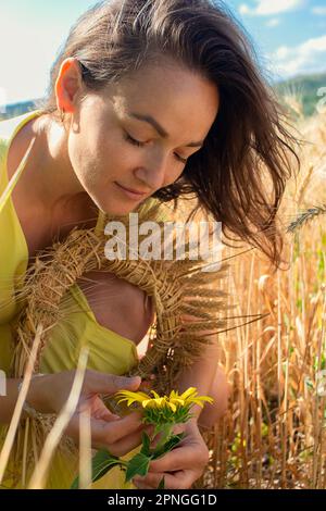 Eine junge Frau in einem gelben Kleid, mit lockerem Haar, kaukasischer Rasse, hockt auf einem Feld, berührt eine gelbe Blume mit den Händen, untersucht sie vorsichtig Stockfoto