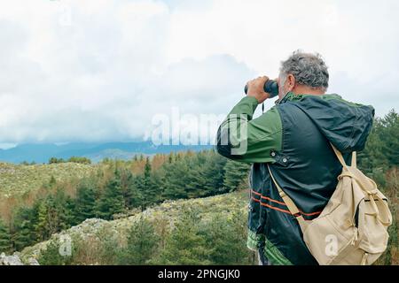 Reifer, bärtiger Reisender, wandert im Bergwald und sieht mit dem Fernglas das Bergtal mit Panoramablick aus. Stockfoto