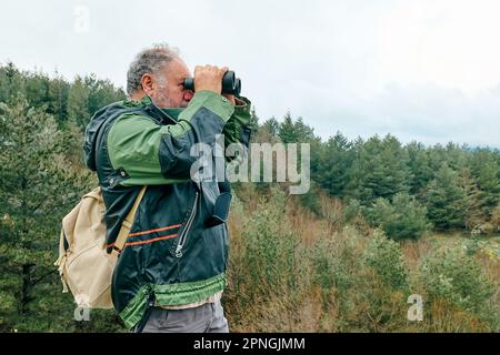 Reifer, bärtiger Reisender, wandert im Bergwald und sieht mit dem Fernglas das Bergtal mit Panoramablick aus. Stockfoto