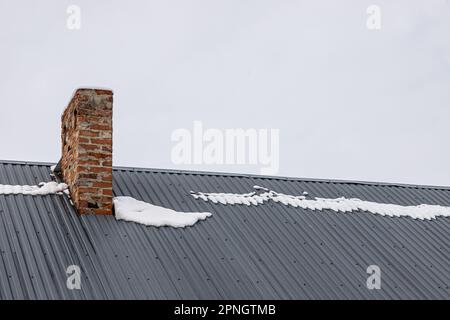 Ein Ziegelkamin auf einem Metalldach im Winter unter dem Schnee. Baukonzept. Stockfoto