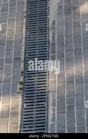 Ein Gitter eines Drainage-Pflastersystems auf einem Fußweg aus quadratischen Steinfliesen, Nahaufnahme eines Regenwasser-Entwässerungssystems. Stockfoto