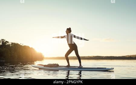 Gleichgewicht ist der Schlüssel. Eine attraktive junge Frau, die im Freien Yoga auf einem Paddelbrett auf einem See macht. Stockfoto