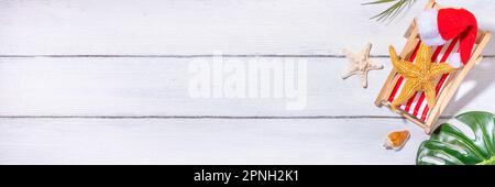 Weihnachten im Juli Sommerferien oder Verkaufshintergrund. Sommerurlaub Accessoires, Muscheln, Seesterne, Klubsessel mit Weihnachtsmannmütze, tropische Blätter. G Stockfoto