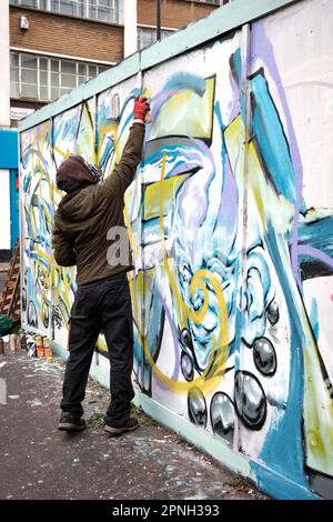 Graffiti-Künstler, der eine Atemmaske trägt, verwendet eine Sprühdose an Bord, um ein Design auf einer Straße in London, Großbritannien, zu malen. Stockfoto