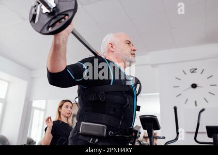 Der ältere Mann im rettungssanitäter-Anzug trainiert mit einem Personal Trainer im Fitnessstudio. Grauhaarige Sportsmann-Squats mit Haarglocke beim Training zur elektrischen Muskelstimulation. A Stockfoto