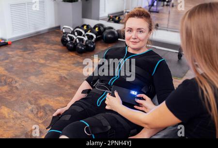 Eine blonde Frau mittleren Alters sitzt auf einem Massagesessel und wartet auf das rettungsdienst-Training im modernen Fitnessstudio. Junge Trainerin stellt notwendige Parameter auf Elektrica Stockfoto