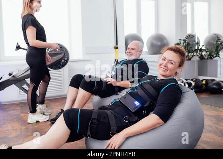 Lächelndes älteres Paar sitzt nach dem rettungsdienst-Training im Fitnessraum auf Massagesesesseln. Junge Trainerin mit Fernbedienung stellt die notwendigen Parameter auf Stockfoto